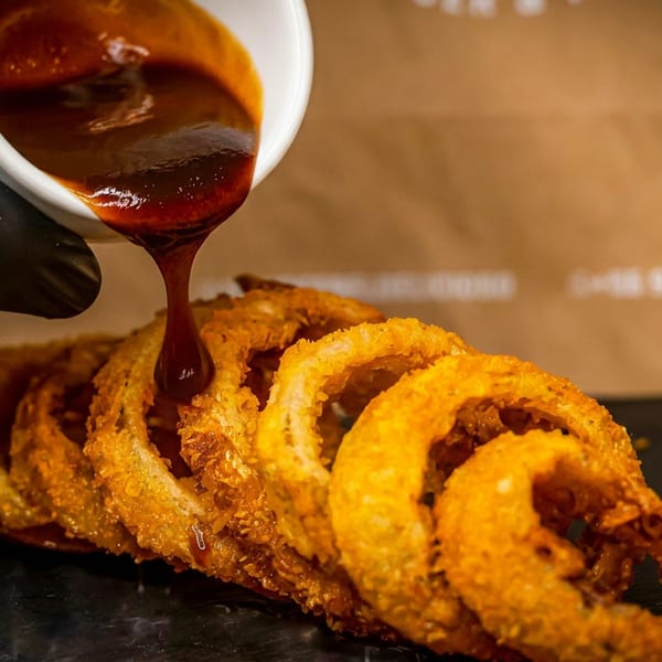 A gloved hand pours rich BBQ sauce over a serving of crispy, golden-brown onion rings.