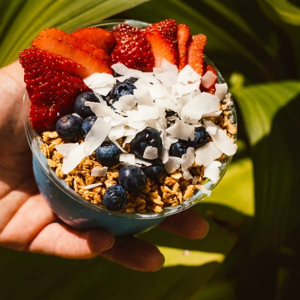 Hand holding a vibrant acai bowl topped with strawberries, blueberries, granola, and coconut flakes.