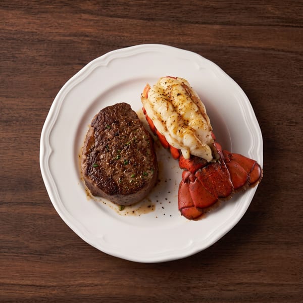Seasoned steak and grilled lobster tail on a white plate over a wooden background.