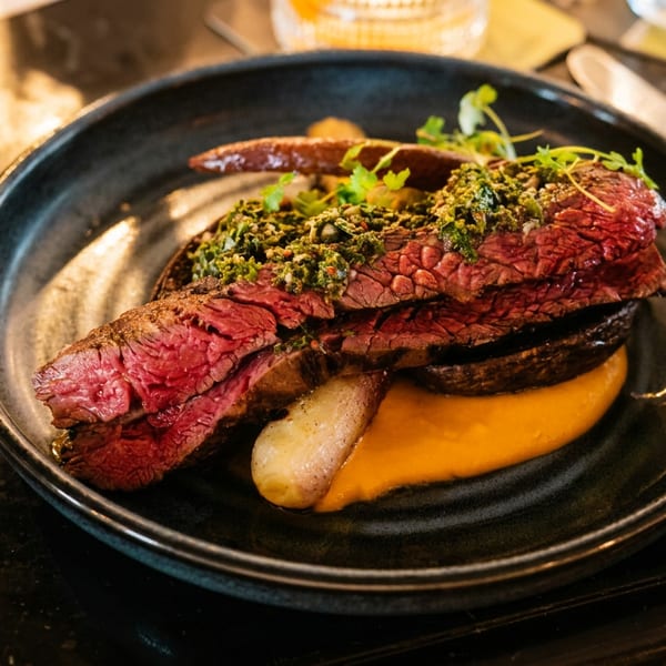 Sliced medium-rare steak with green herb crust, orange puree, and roasted vegetables on a dark plate.